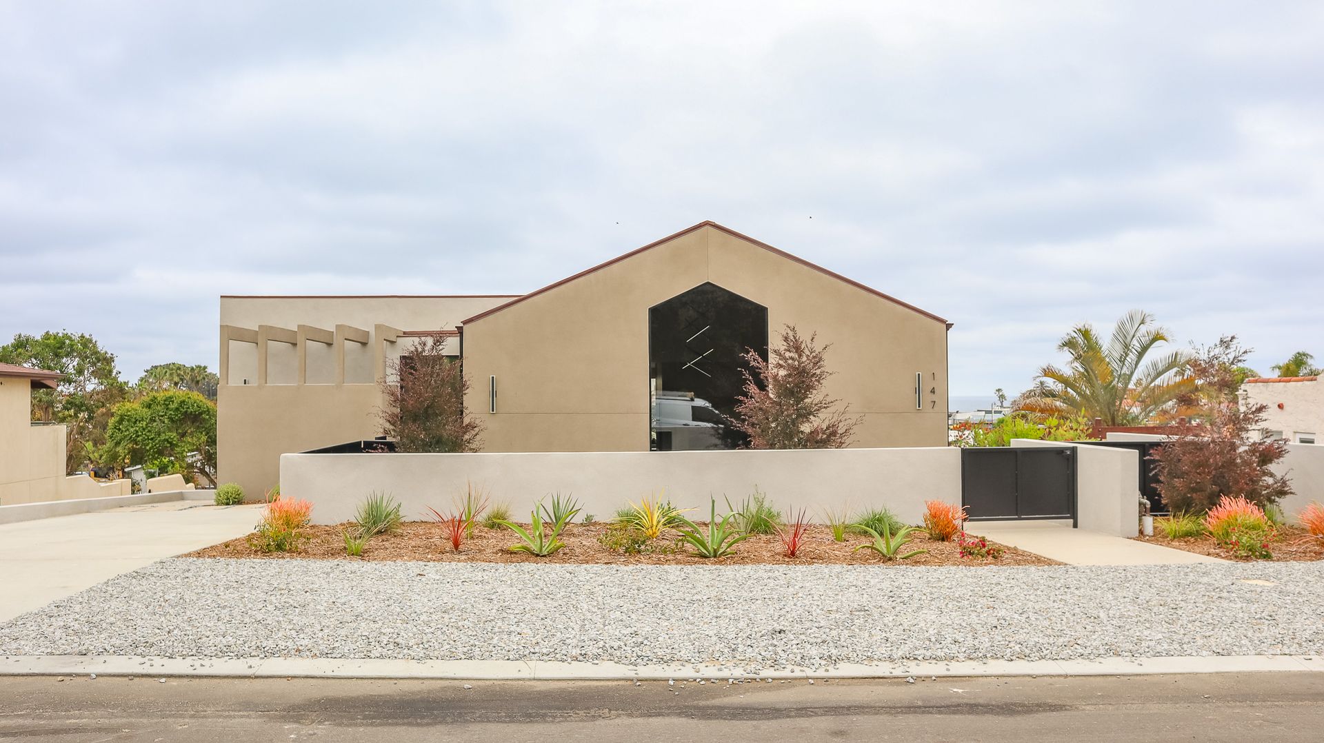 Beige modern house with a gabled roof and a low concrete wall with landscaping.