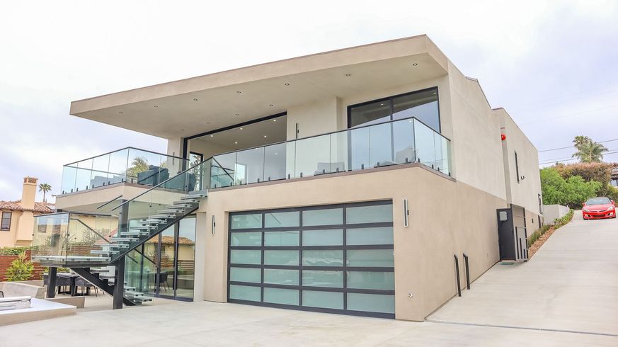 A tan, two-story modern house with a glass-railed balcony, a large frosted glass garage door, and a metal spiral staircase.