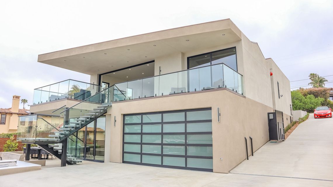 A tan, two-story modern house with a glass-railed balcony, a large frosted glass garage door, and a metal spiral staircase.