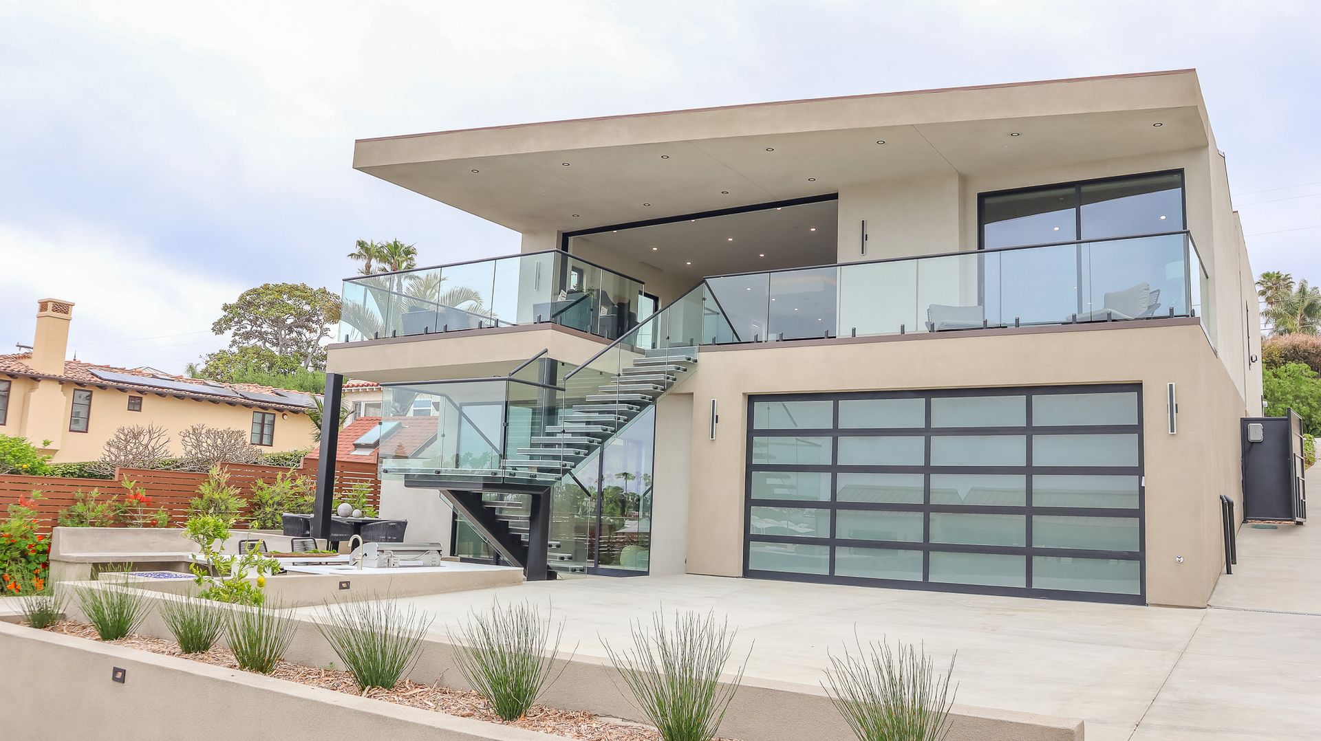 Modern two-story house with glass balcony and garage doors, stairs, and landscaping.