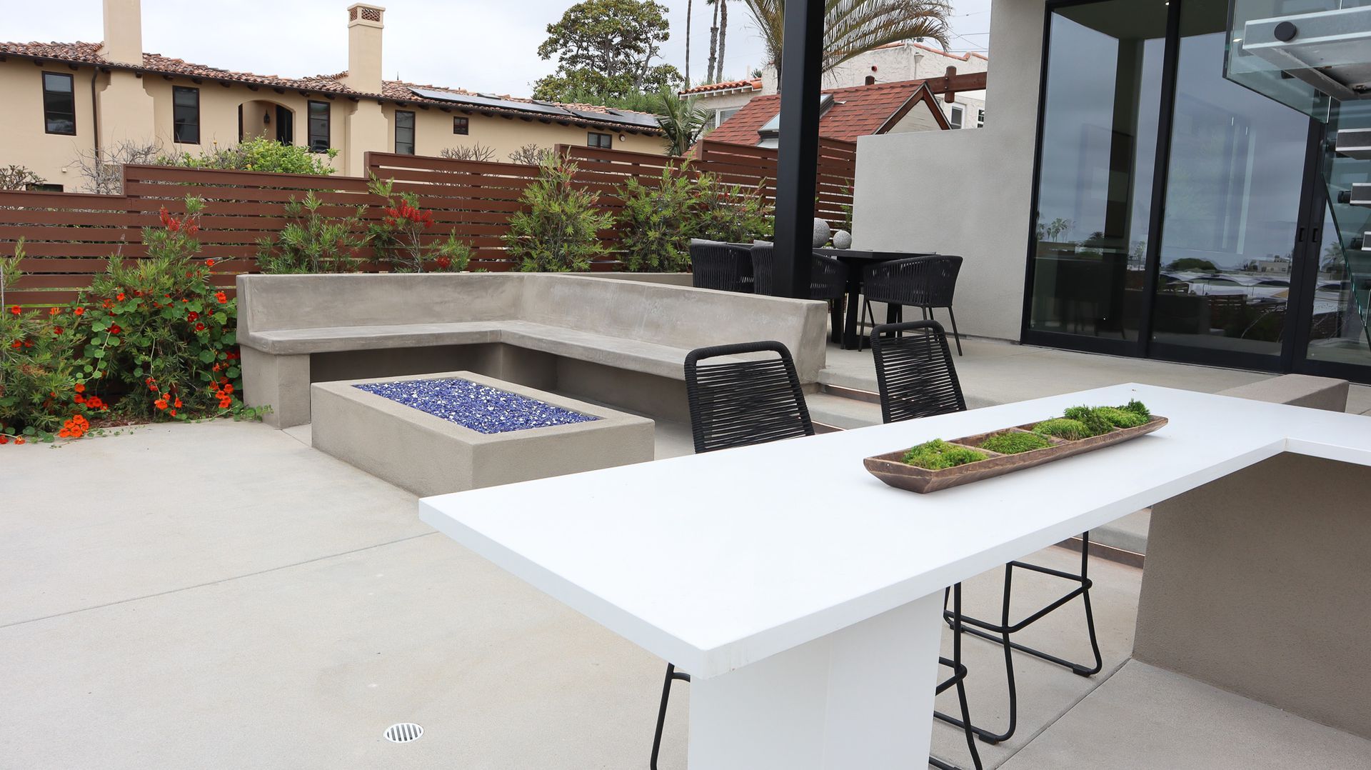 Patio with concrete benches, fire pit, and white bar. Black chairs and greenery are present.