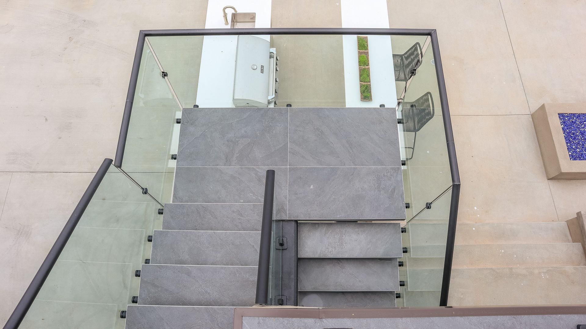 Staircase with glass railing, gray tile steps, and metal handrail. Beige walls.