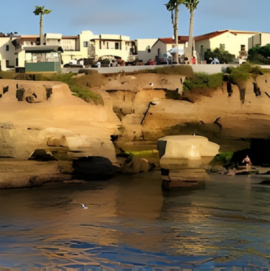 Sunlight hits tan cliffs above a calm bay with buildings and palm trees in the background under a blue sky.