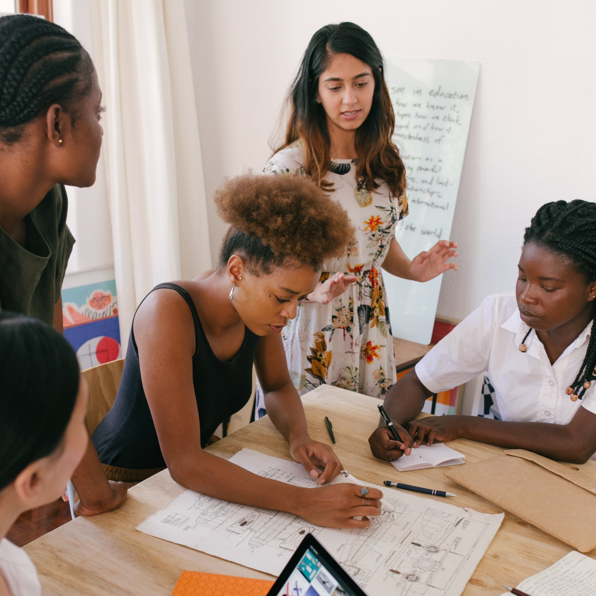 Un grupo diverso de estudiantes colabora alrededor de una mesa de madera, trabajando en papeles y un dibujo para un proyecto.