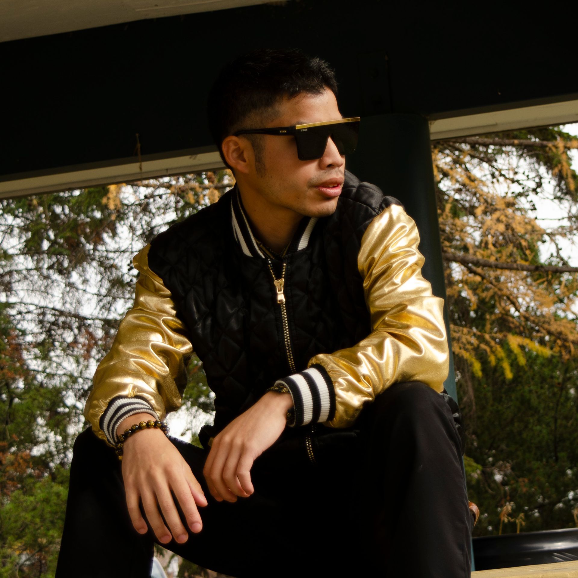 a pensive looking man of thai complexion in a flamboyant black & gold letterman jacket wearing sunglasses