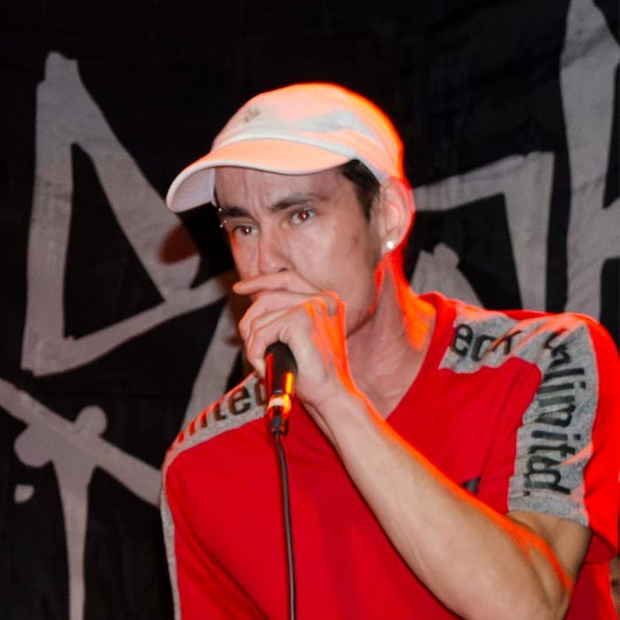 A pale caucasian man holding a microphone with a red shirt & white baseball cap