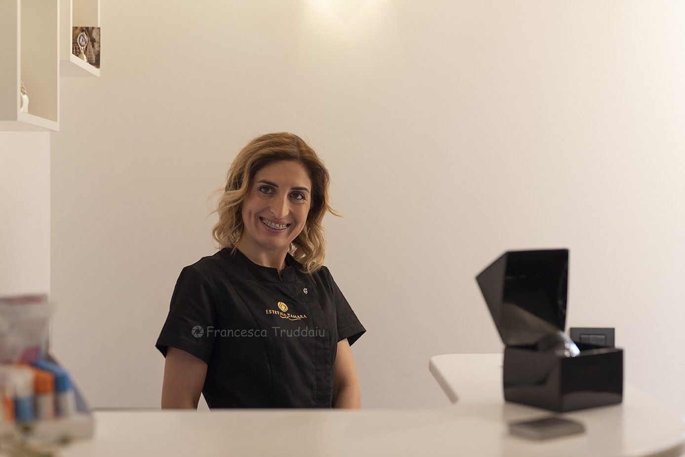 Donna sorridente dietro un bancone, con indosso una maglietta nera, pareti bianche sullo sfondo, scatola nera sul bancone.