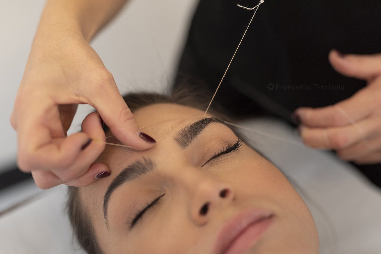 Donna che si fa truccare le sopracciglia con il filo. Primo piano delle mani che le passano il filo sulle sopracciglia, con gli occhi chiusi.