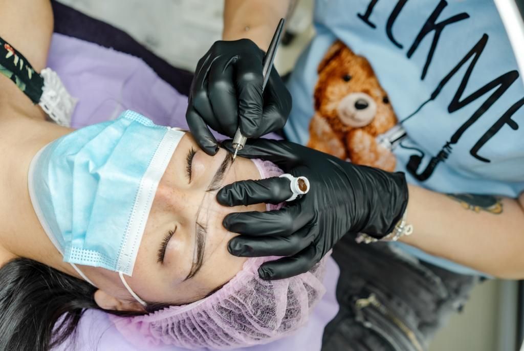 Persona sottoposta a microblading delle sopracciglia da un professionista. Mani guantate di nero che tengono uno strumento sul viso del paziente.