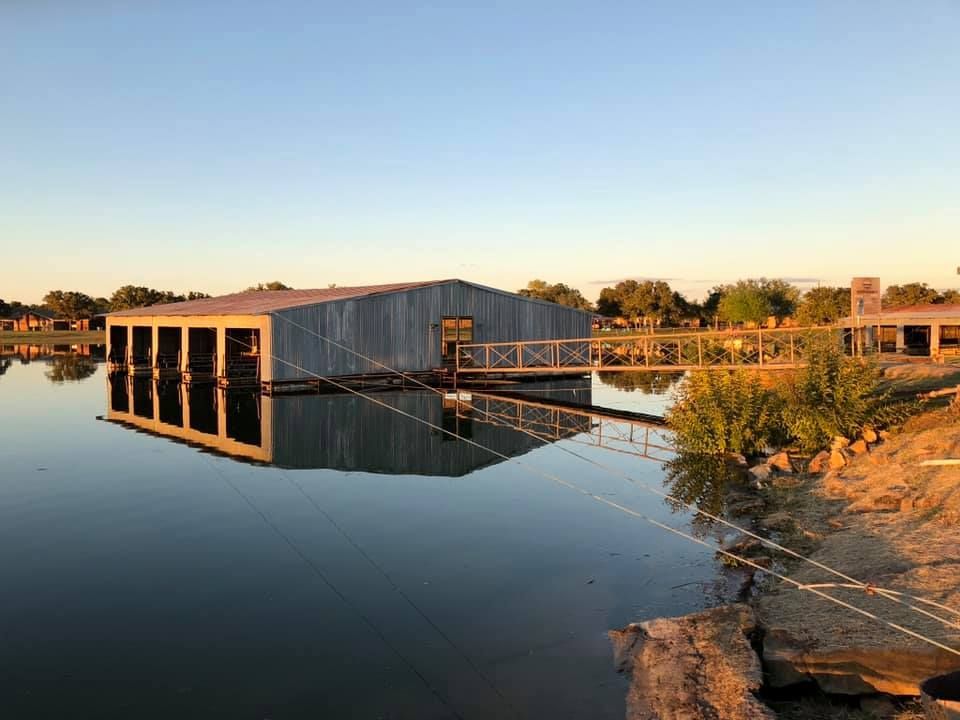 A floating boathouse on a calm lake at sunset. The building has multiple open boat slips and is reflected in the water.