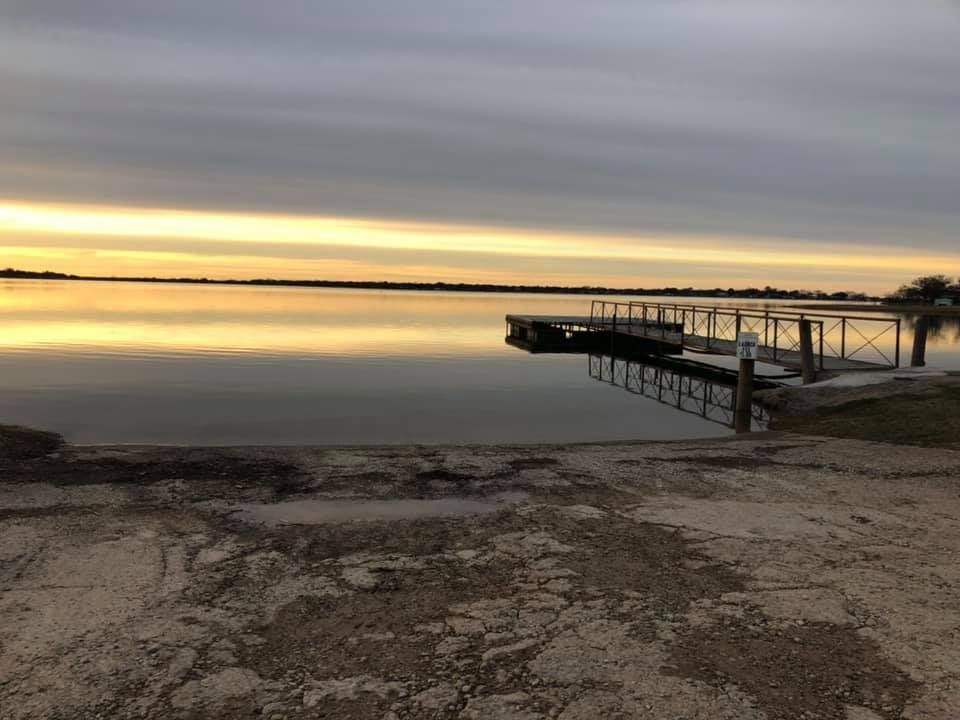 Calm lake with a dock under a cloudy sunset sky, the water reflecting the golden light. A boat launch is in the foreground.