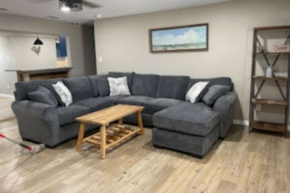 A sectional sofa with an ottoman and coffee table in a living room with light wood flooring and a wooden bookshelf.