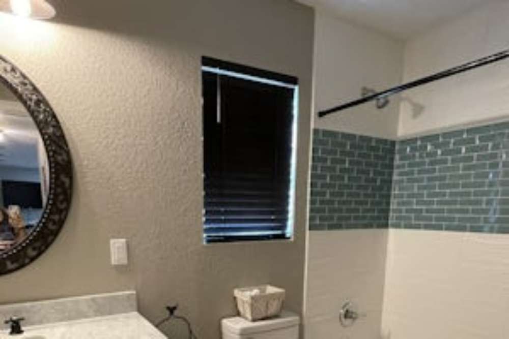 Bathroom interior: a round mirror, window with dark blinds, and a tiled shower with a gray-green brick pattern.