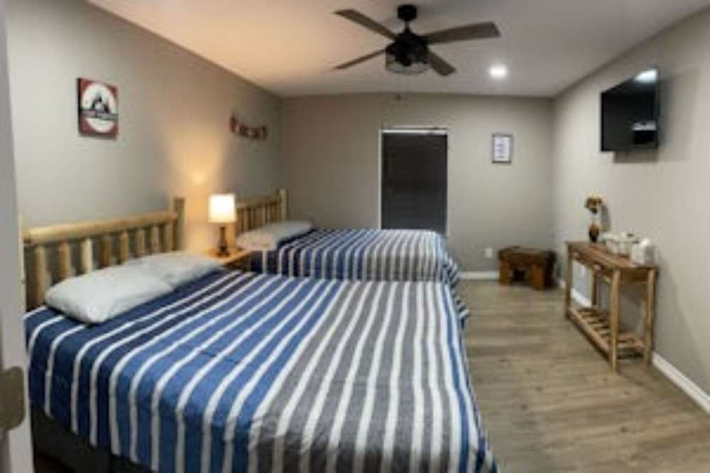 A hotel room featuring two queen beds with blue and white striped comforters. A TV, lamp, and small table are also visible.