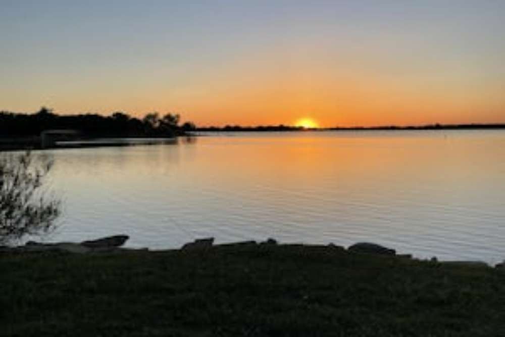 Sunset over a calm lake; orange and gold hues reflect on the water. Silhouetted trees line the horizon.