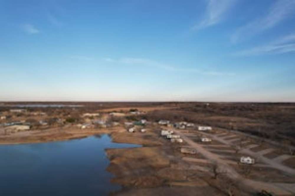 Aerial view of a campground by a lake under a blue sky. RVs are parked along dirt roads near the water's edge.