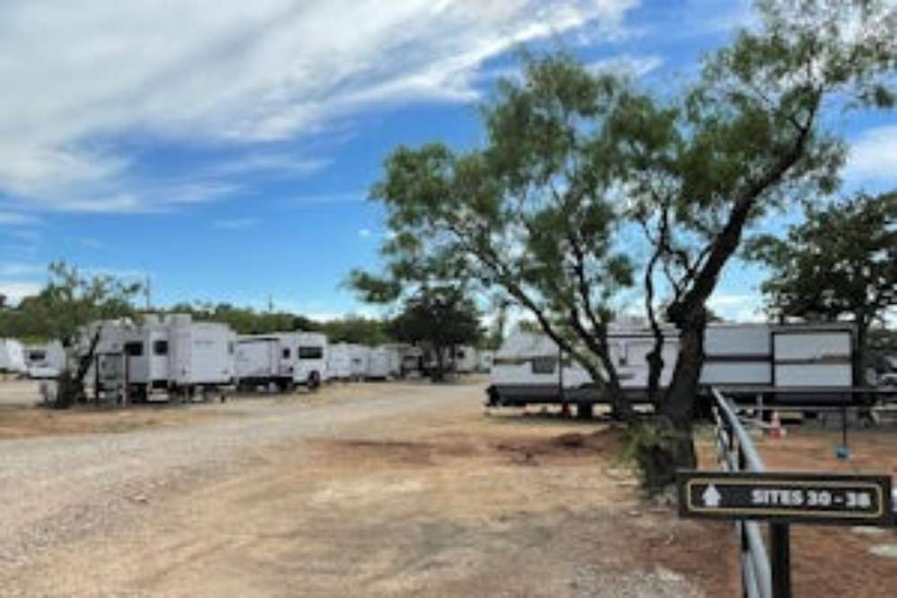 RV campground with several white RVs parked on gravel lots under a blue sky with scattered trees. Sign points to sites 30-36.