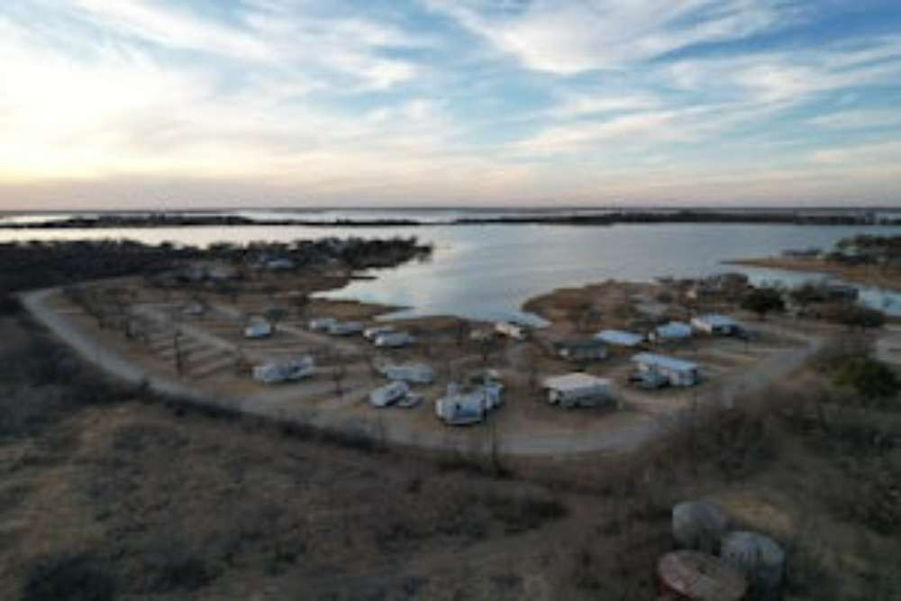 An aerial view of a lakeside RV park under a cloudy sky. RVs are parked near the water, with some trees and brush in the foreground.