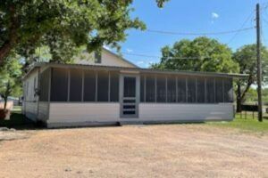 A white building with a screened porch, in front of a gravel driveway and trees. The sky is blue.