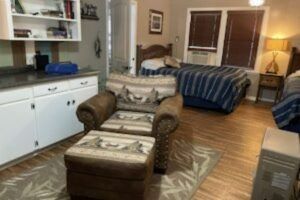 Cozy living space with a patterned armchair and ottoman, two beds with blue bedding, and a small kitchen area.