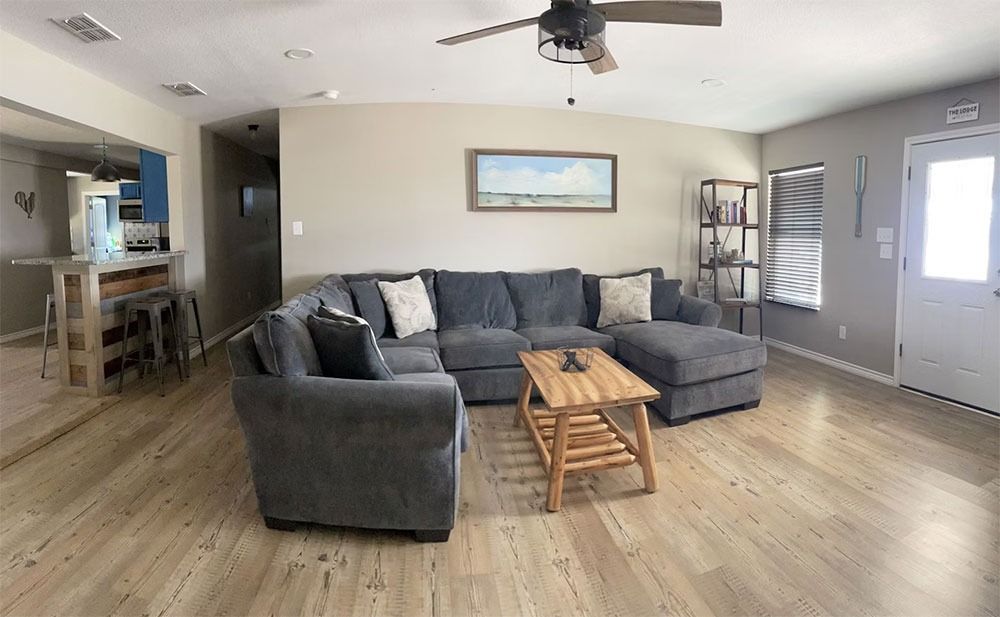 Spacious living room with gray sectional sofa, wooden coffee table, and light wood floors. A painting hangs above the couch and a door is on the right.