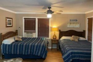 A hotel room with two queen beds covered in striped bedding.  A lamp sits on a nightstand between the beds, and a ceiling fan hangs overhead.