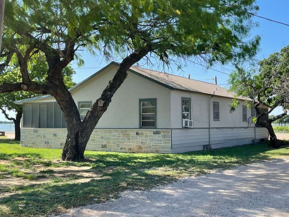 A single-story house with a stone base and light-colored siding, partially shaded by trees, sits on a grassy lot.