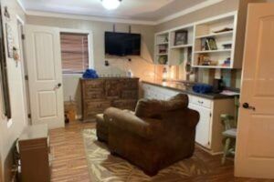 A small room with hardwood floors, an armchair, built-in shelves, and a TV. There's a door on the left, and a cabinet is on the right.