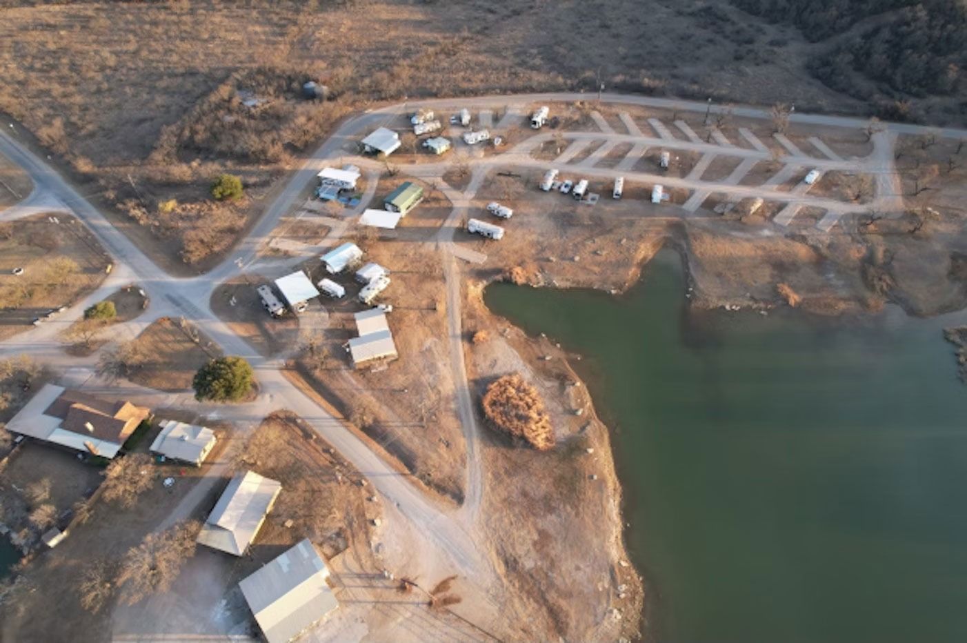 Aerial view of a campsite with RVs and cabins near a lake; brown and green landscape.