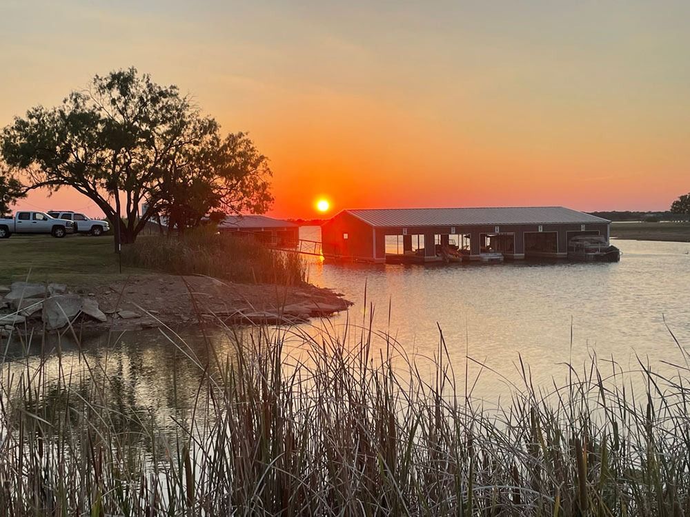 Sunset over a lake with a boathouse, tall grasses, and a tree. Golden light reflects on the water.