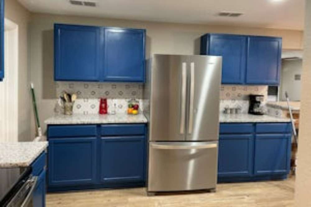 A kitchen with blue cabinets, stainless steel refrigerator, and white countertops with gray speckles.