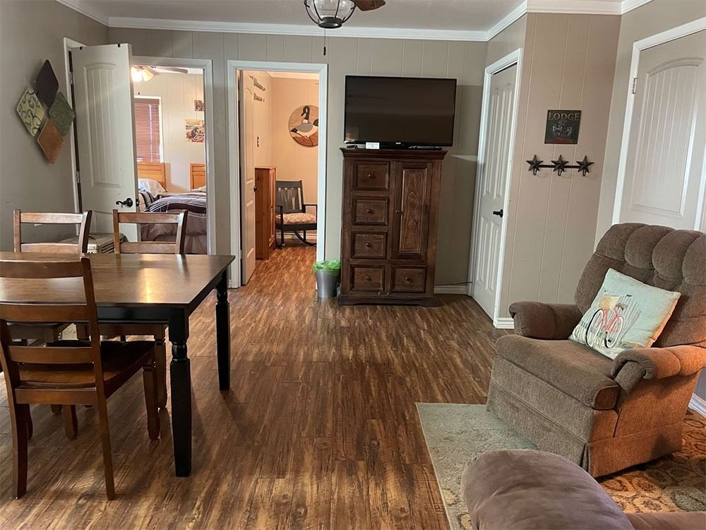 A living room and dining area with a wooden table, chairs, and a recliner. A television sits atop a decorative cabinet.