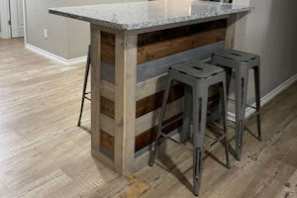 A kitchen island with a granite countertop, wooden paneling, and two gray bar stools. The island sits on a light wood floor.