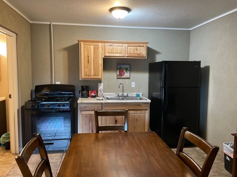 Small kitchen with a wooden table and chairs, stove, sink, and black refrigerator. Light wood cabinets and neutral walls.