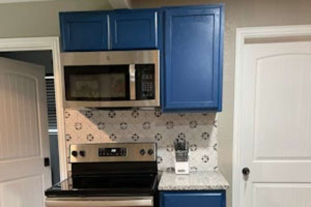 Blue kitchen cabinets with a stainless steel microwave above a matching range, set against a patterned backsplash and white doors.