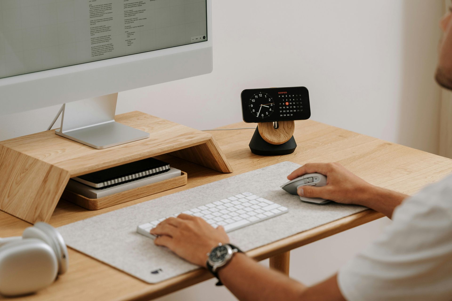 Person at wooden desk using a computer and peripherals, with a phone charging on a stand.