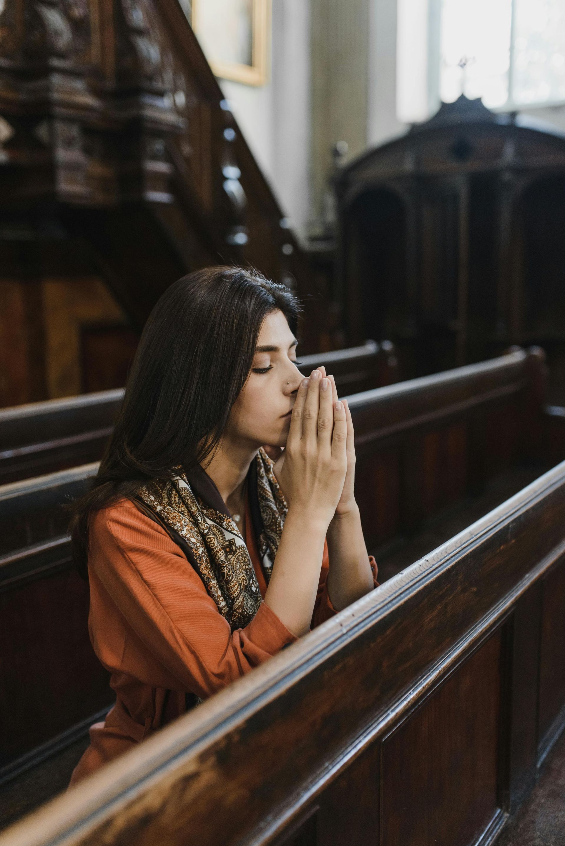 Woman in orange dress praying in a church pew with hands clasped, eyes closed.