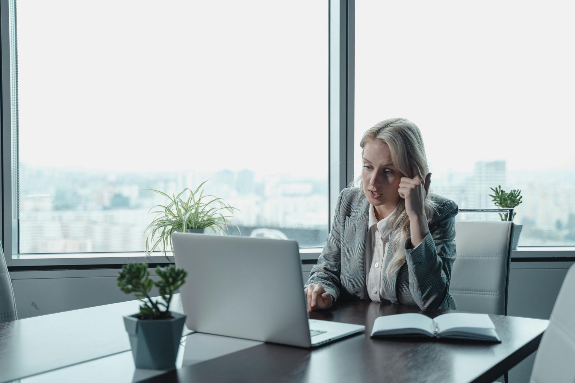 Woman in a blazer sits at a desk with a laptop, looking thoughtful. Office with city view.