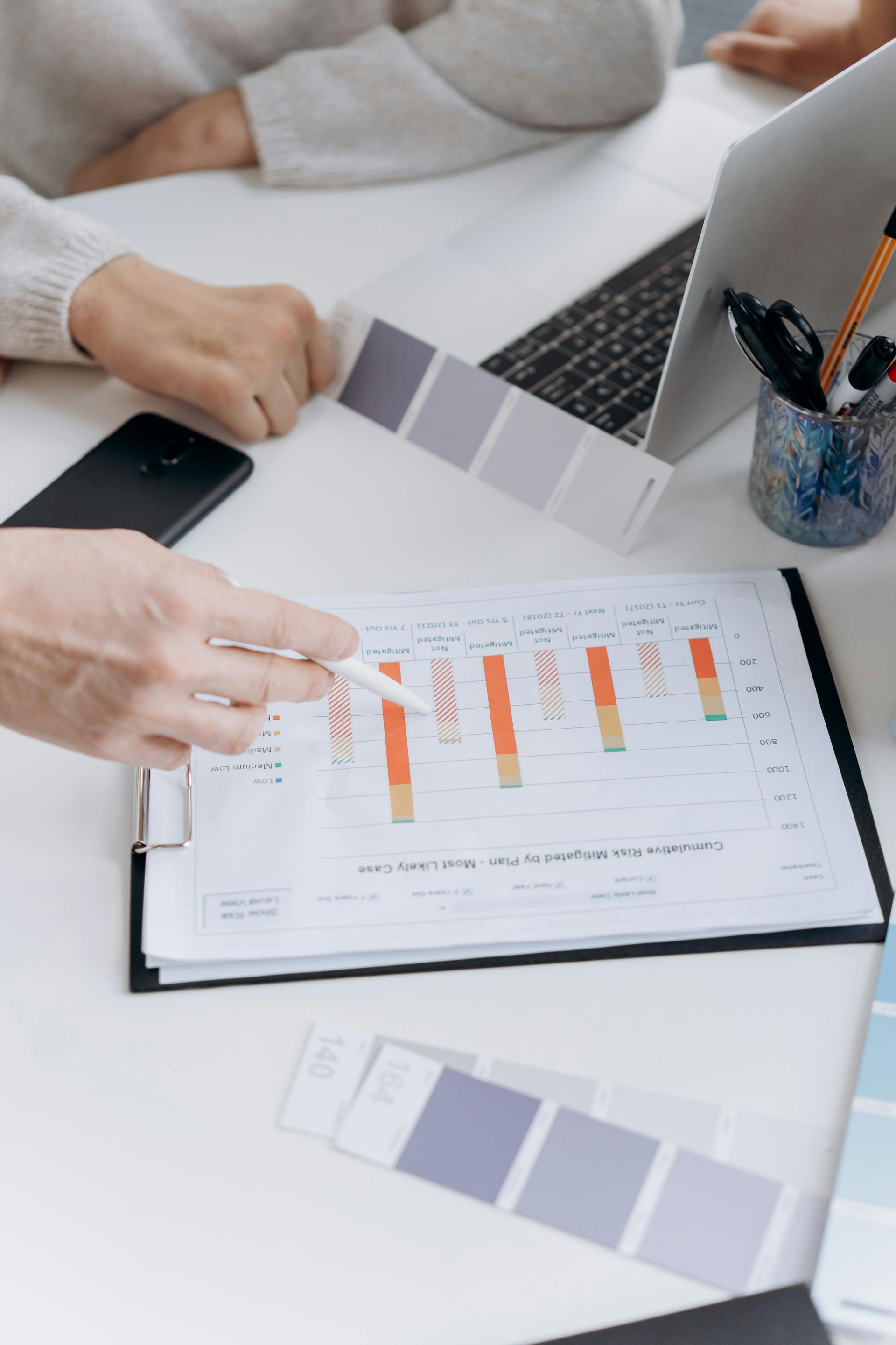Hands pointing at a bar graph, color swatches, laptop, and phone on a white table.