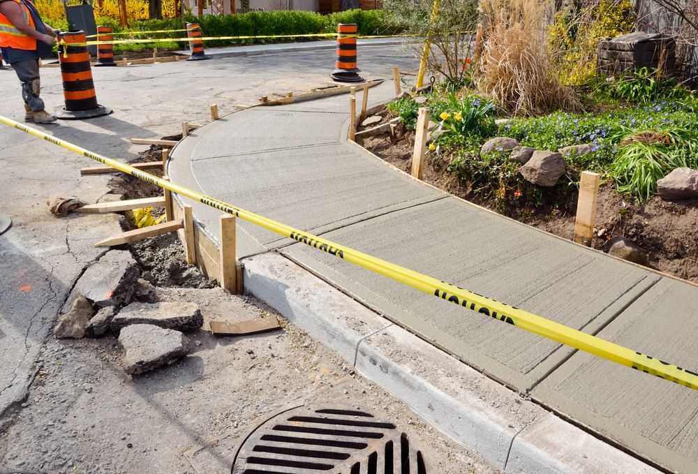 New concrete sidewalk under construction, blocked by caution tape; curb, street, and drain visible.