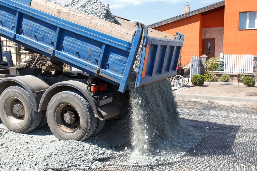 Dump truck dumping gravel on a road in front of a house.