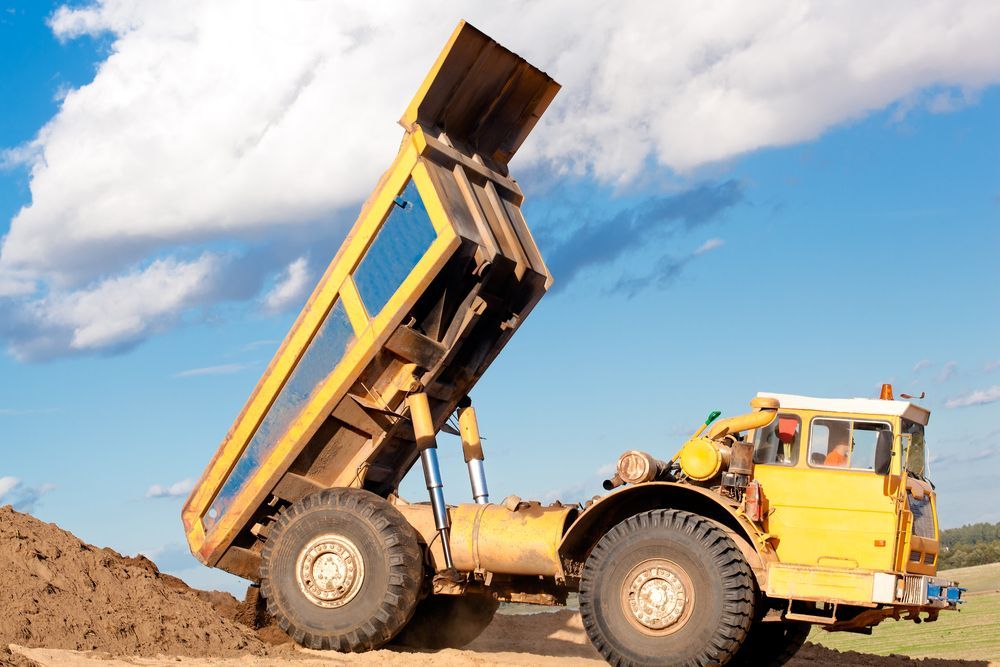 Yellow dump truck, bed raised, unloading dirt against a blue sky.