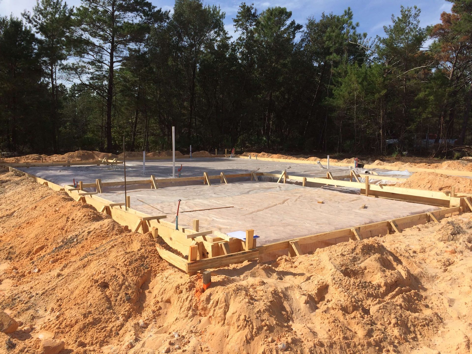 A rectangular concrete foundation with wooden frame, in a sandy construction site with trees in the background.