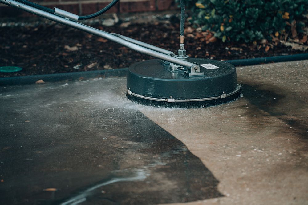 A person pressure washing a concrete surface with a circular cleaning head; water sprays outwards.