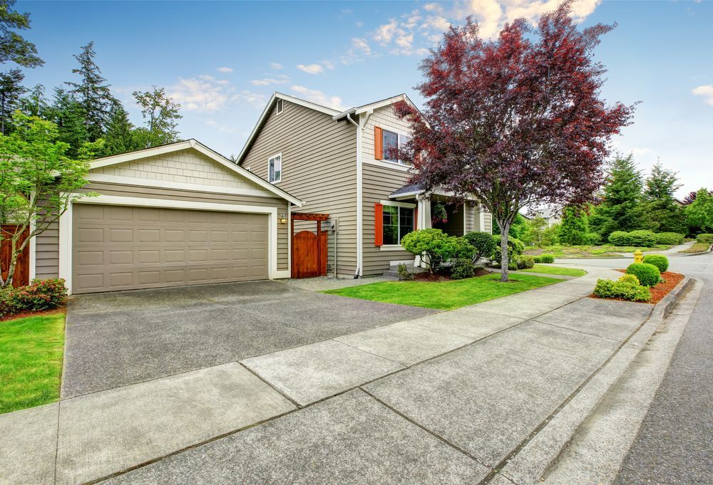House with attached garage, driveway, and sidewalk. Red tree, shrubs, and green lawn. Blue sky.