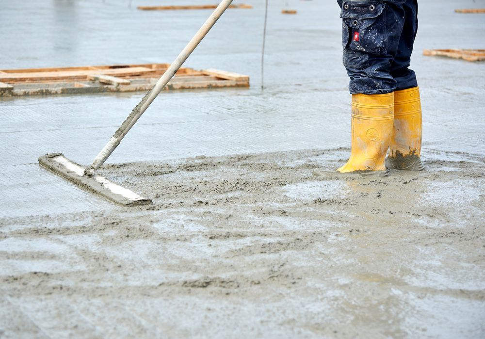 Person smoothing wet concrete with a screed; wearing yellow boots, construction site.