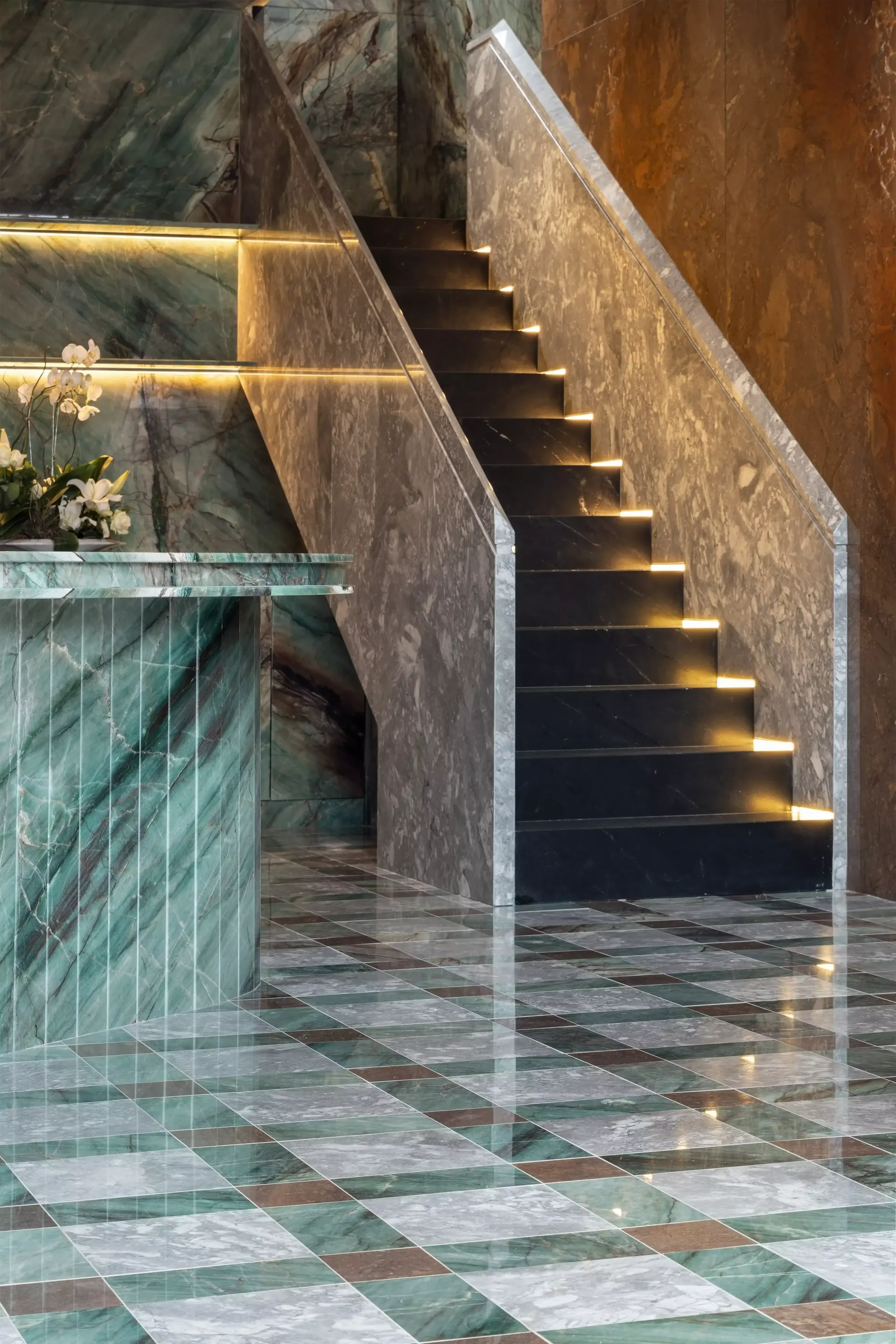 Hall de entrada elegante com paredes e escadaria de mármore, degraus iluminados e piso em ladrilhos verdes e marrons.
