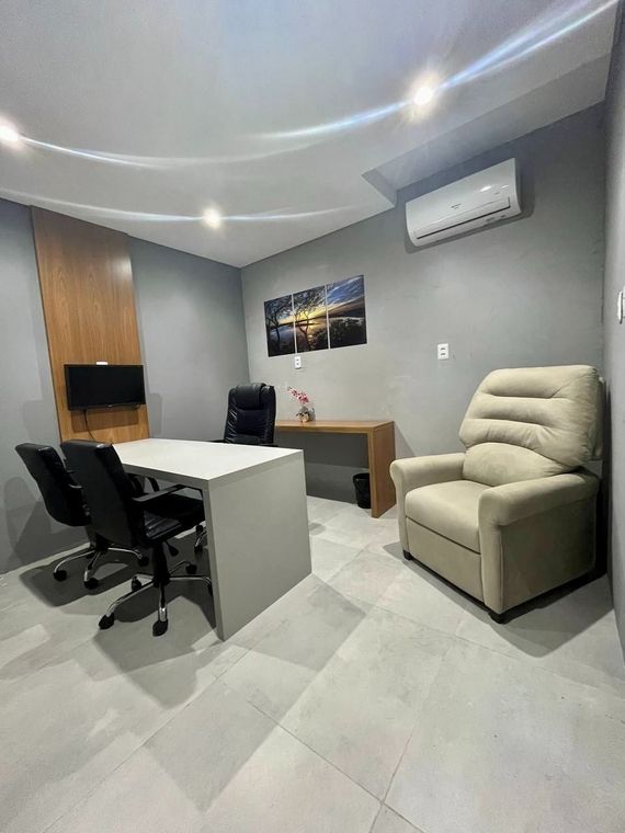 Escritório com mesa, cadeiras e poltrona reclinável. As paredes são cinza e há uma TV, ar-condicionado e obras de arte.