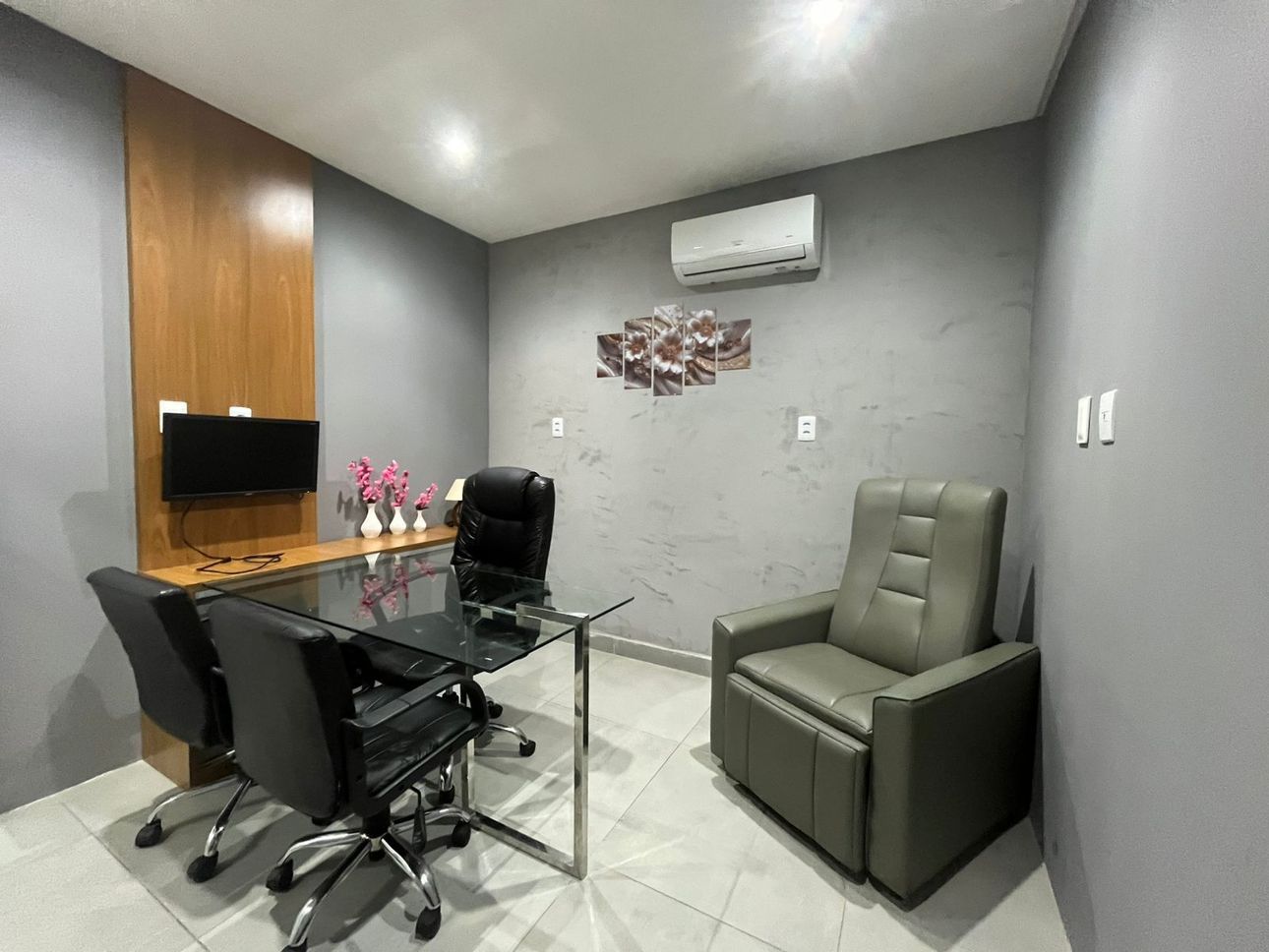 Escritório com mesa de vidro, cadeiras pretas, paredes cinza e uma poltrona reclinável de couro.