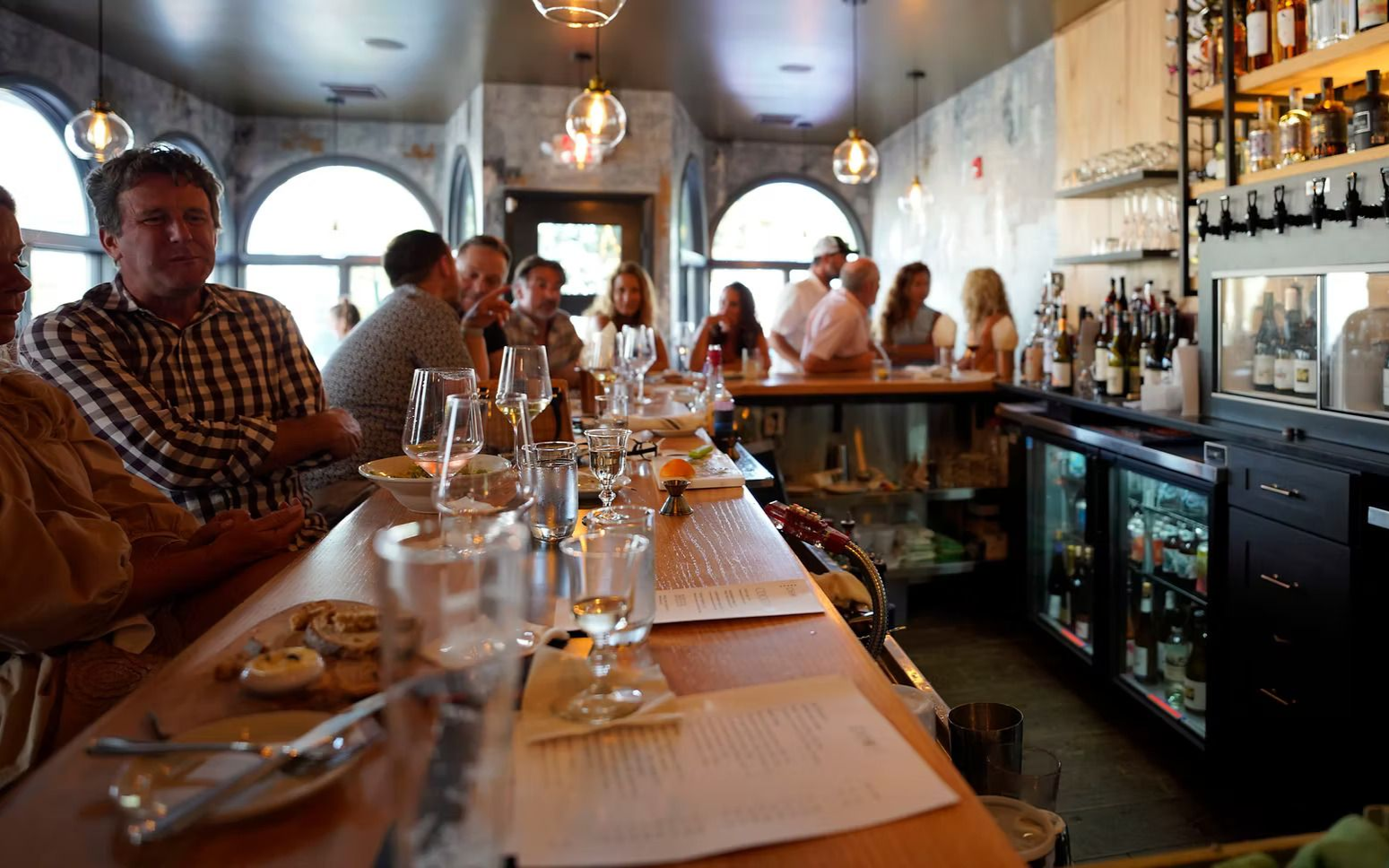 People at a bar, enjoying drinks. Bottles and glasses on the bar, bright interior.